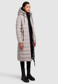 Gaišpelēkā pūkaini jaka ar kapuci, kurai ir quilted dizains, ar rāvējslēdzēja sānu kabatām un gludu faktūru. Nēsā virs melnas apģērba un zābakiem.