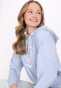 Světle modrá mikina s kapucí vyrobená z měkké tkaniny, s barevným kruhovým grafickým designem na přední straně, bílými šňůrkami a žebrovanými manžetami.