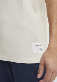 Kremfarget bomulls-T-skjorte med et rektangulært merke med "11 PROJECT" nær kanten, som viser en myk tekstur og et enkelt design.