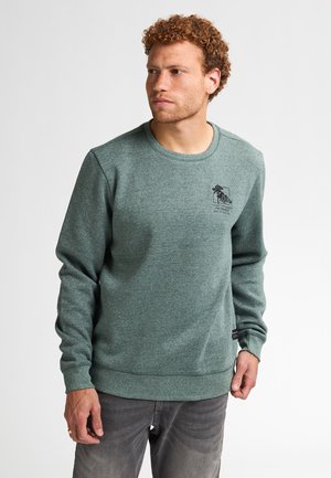 Sweatshirt vert en tissu texturé, avec un col rond, des poignets côtelés et un logo imprimé sur la poitrine. Porté sur un jean gris.