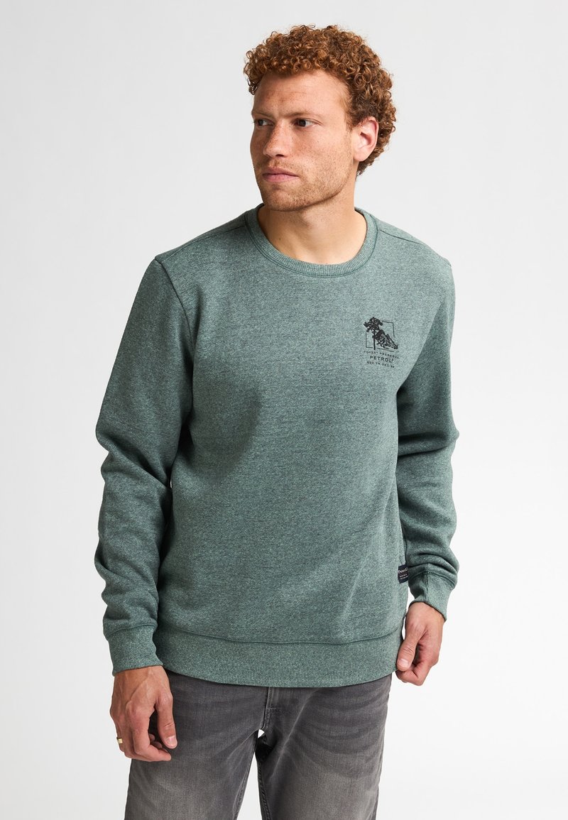 Felpa verde realizzata in tessuto testurizzato, con colletto a girocollo, polsini a costine e logo stampato sul petto. Indossata sopra jeans grigi.