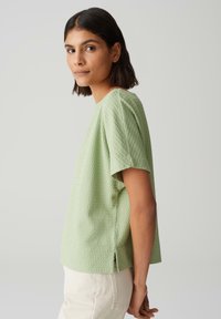 Haut vert clair à manches courtes en tissu texturé, encolure ronde et fentes latérales. Porté avec un pantalon blanc cassé, mettant en avant une coupe décontractée.