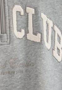 Sudadera gris con letras de felpa en color marfil que dicen "CLUB" y texto cursivo "Establecido 2012", con un cuello de media cremallera.