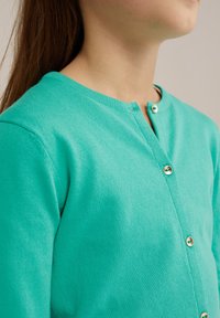 Tirkīza krāsas adīta žakete ar apaļu dekoltē, kas rotāta ar zelta toņu pogām gar apģērba priekšējo daļu un ar mīkstu, gludu tekstūru.