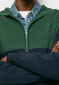 Grön och marinblå zip-up hoodie med ribbad textur, justerbar huva och logotypdetalj på ärmen. Lagd över en denimskjorta.
