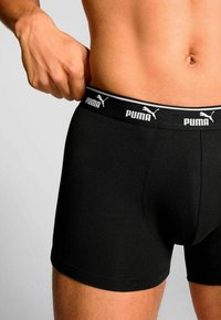 Man adjusting waistband of black Puma boxer briefs, showing torso and upper thighs against a light background.