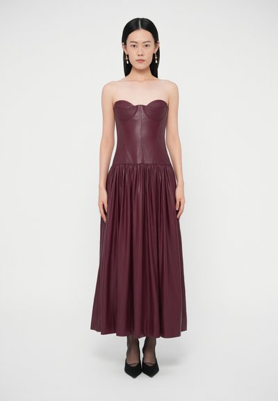 Weinrotes trägerloses Kleid aus Leder mit figurbetontem Oberteil und gerafftem Rock. Glatte Textur mit dezentem Glanz, kombiniert mit schwarzen High Heels.
