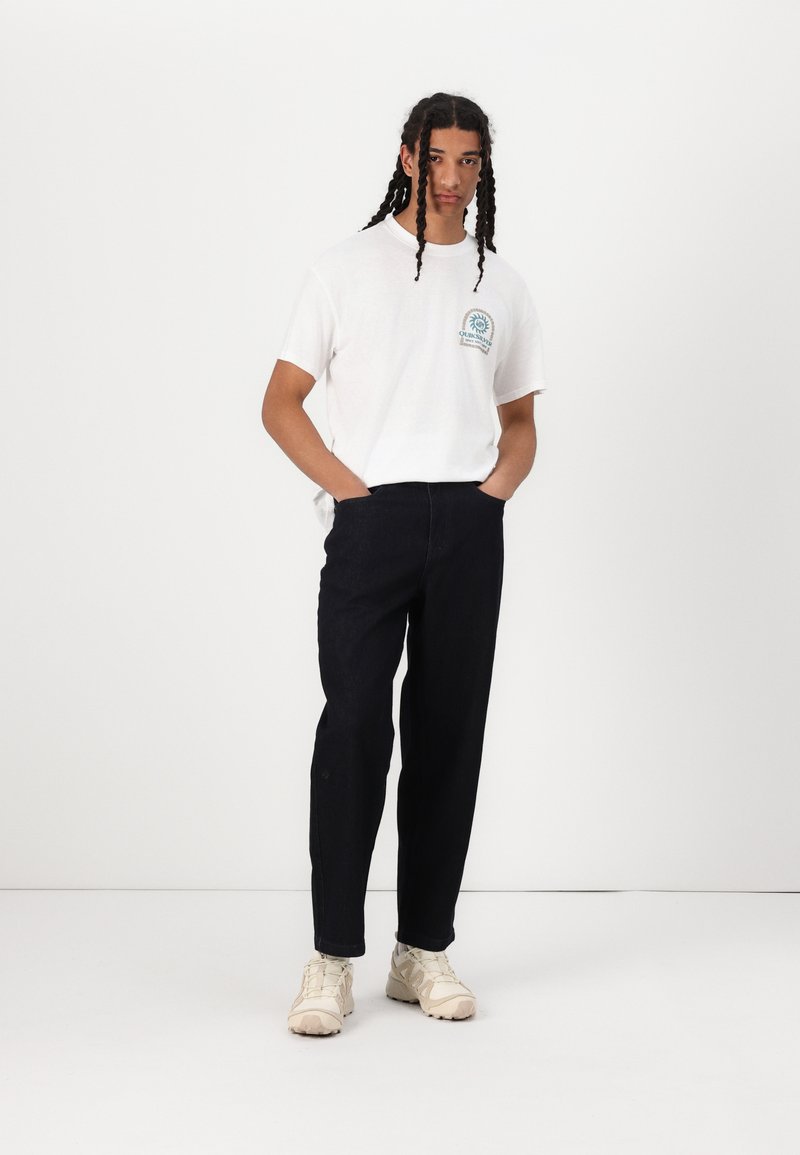 T-shirt bianca di cotone con un logo grafico blu sul petto, abbinata a pantaloni in denim blu scuro e sneakers beige, il tutto su uno sfondo neutro.