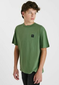 T-shirt en coton vert à manches courtes, coupe décontractée, avec un patch logo noir carré sur la poitrine gauche. Ourlet plat et design à col rond.