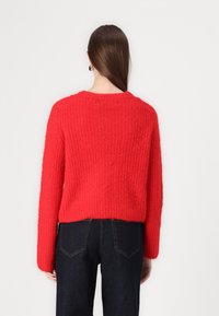 Pull en tricot rouge texturé avec un col rond et des manches longues, associé à un jean bleu foncé. La vue de dos montre une coupe décontractée.