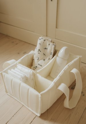 Organizzatore morbido in tessuto con scomparti che contengono panni piegati e un biberon, posizionato su un pavimento in legno vicino a un armadietto.