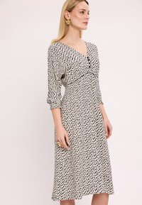 Czarna i biała sukienka w wzory z dekoltem w kształcie litery V, długimi rękawami i guzikami na biuście. Tkanina jest miękka i ma zwiewny fason.