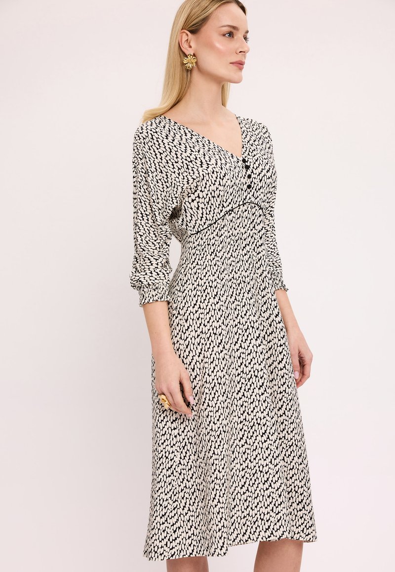 Czarna i biała sukienka w wzory z dekoltem w kształcie litery V, długimi rękawami i guzikami na biuście. Tkanina jest miękka i ma zwiewny fason.