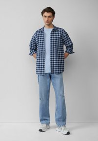 Gezicht op een blauw-wit geblokt shirt dat over een lichtblauw T-shirt is gedragen, gecombineerd met gescheurde lichtblauwe jeans en grijze sneakers. Eenvoudig ontwerp.