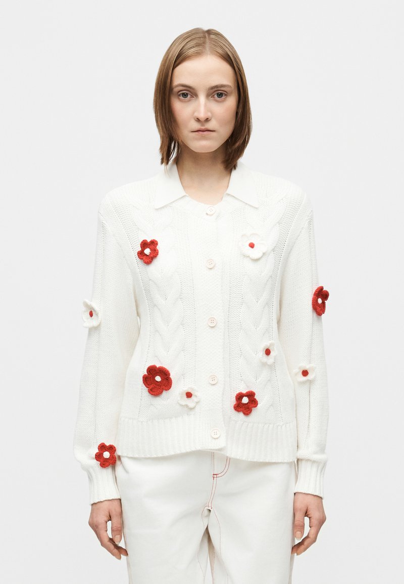 Hvit strikket cardigan med kabelstrikk-mønster, blomsterapplikasjoner i rødt og hvitt, og krage, lukket med lyse knapper.