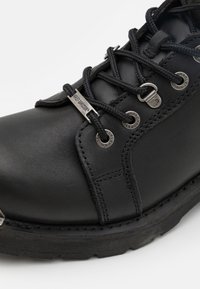 Bota de cuero negro con cordones, puntera redondeada, herrajes plateados y suela texturizada. Presenta detalles de costura y acentos de marca en relieve.