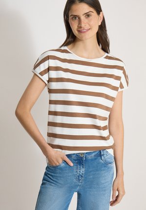 Femme aux cheveux bruns portant un haut rayé blanc et marron à manches courtes et un jean bleu, debout avec une main dans la poche.