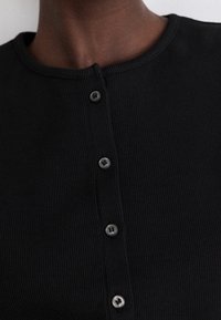 Schwarzes, geripptes Oberteil mit rundem Halsausschnitt und fünf vorne angebrachten Knöpfen in passender Farbe. Der Stoff hat eine strukturierte Optik.