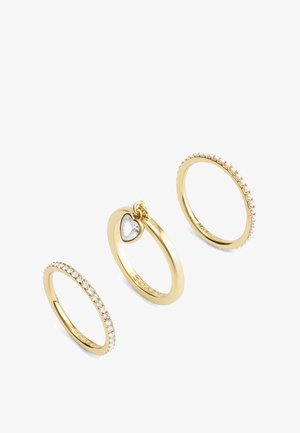 Trois bagues en or aux designs différents : une avec de petites perles, une avec des pierres transparentes, et une avec un pendentif en forme de cœur.