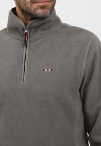 Pilkas vilnonis golfas su aukštu apykakle, ketvirtiniu užtrauktuku ir išsiuvinėtu logotipu raudona, balta ir mėlyna spalvomis, turintis tekstūruotą paviršių.