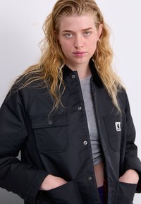 Schwarze wasserdichte Jacke mit Druckknopfverschluss, zwei Fronttaschen und einem Logopatch auf der linken Brust. Glatte Textur und lässige Passform.