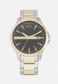 Armani Exchange pulkstenis ar melnu teksturētu ciparnīcu, zelta akcentiem, sudraba un zelta nerūsējošā tērauda siksnu un datuma displeju pie 3 stundām.