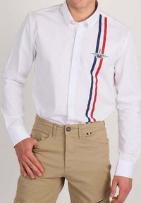 Homme portant une chemise blanche à boutons avec des rayures verticales rouges et bleues et un emblème d'avion, associée à un pantalon beige avec un petit détail tricolore.