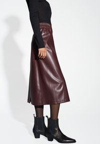 Bordeauxrote Leder-Midirock mit einem elastischen Bund, kombiniert mit schwarzen Stiefeletten mit spitzer Zehe und niedrigem Blockabsatz.