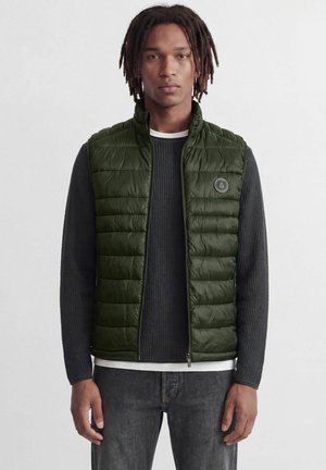 Ung mand iført en åben mørkegrøn vatteret vest over en sort strikket sweater og mørke jeans, stående mod en ensfarvet hvid baggrund.