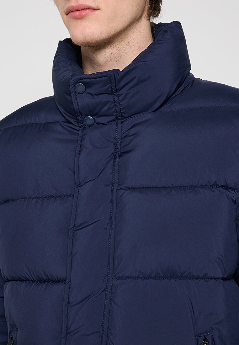 Granatowa, pikowana kurtka z wysokim kołnierzem, z dwoma zatrzaskami i gładką, lekko błyszczącą fakturą. Zawiera poziome przeszycia.