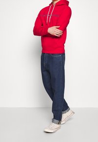 Sudadera roja con cordones blancos y logo de texto, combinada con jeans azul oscuro y zapatillas blancas con acentos rojos. Diseño de atuendo casual.