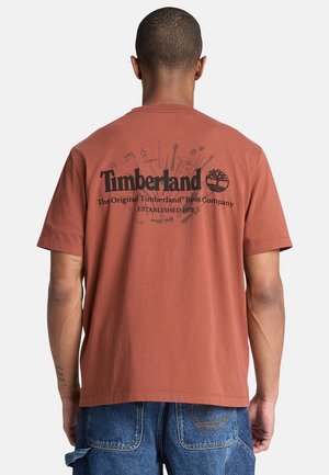 Mann trägt ein rostfarbenes Timberland-T-Shirt mit Logo und Schriftzug auf dem Rücken, kombiniert mit blauen Jeanshosen mit sichtbaren Nähten an den Taschen.