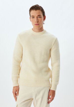 Cream-colored knit sweater with a ribbed texture, round neckline, and fitted cuffs. Worn over light beige pants. Simple design, no patterns.