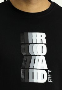 Maglietta nera in cotone con tipografia audace e stratificata in argento e bianco. Il testo recita "La strada non è un luogo, è un sentimento."