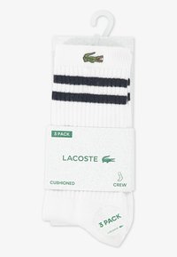 Chaussettes blanches montantes en lot de trois, avec deux bandes horizontales noires et un logo crocodile vert brodé sur chaque chaussette.