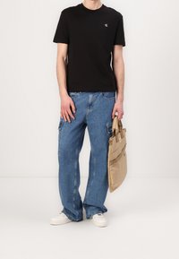 Persona vestida con una camiseta negra de Calvin Klein, vaqueros cargo azules holgados, zapatillas blancas, sosteniendo una bolsa de tote beige frente a un fondo sencillo.