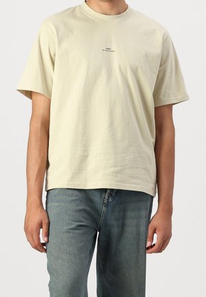 Homme portant un T-shirt beige uni à manches courtes avec un petit texte noir sur la poitrine et un jean en denim bleu, debout devant un fond uni.