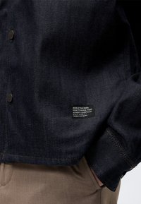Veste en denim foncé avec des boutons noirs et une petite étiquette rectangulaire sur le bas devant au-dessus d’un pantalon beige avec la main dans la poche.