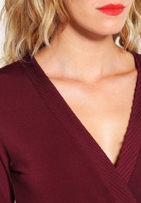 Burgundy omslagstopp med texturerad, ribbad v-ringning och slät tyg. Halsringningen har en djup skärning som visar hud och nyckelben.