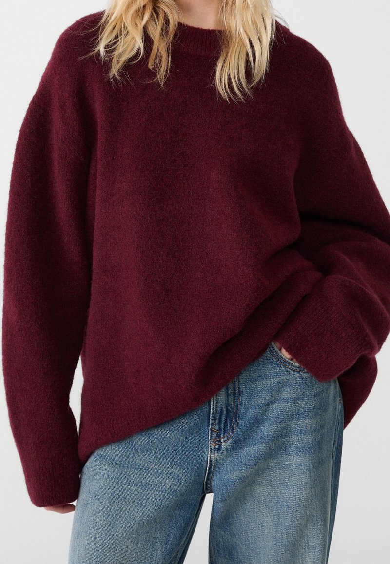 Personne portant un pull bordeaux ample à manches longues avec un jean bleu, une main dans la poche, et des cheveux blonds partiellement visibles.