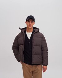 Jachetă puf premium maro cu textură matlasată, închidere cu fermoar și glugă, purtată peste un pulover negru și pantaloni bej, pe un fundal alb.