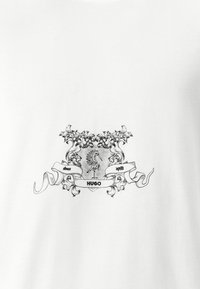 T-shirt en coton blanc avec un design graphique noir représentant un emblème héraldique avec des détails complexes, un coq, et le texte "HUGO" et "depuis 1993."