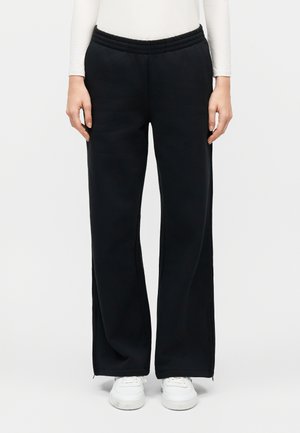 Άτομο που φοράει μαύρο παντελόνι με φαρδιά γραμμή και ελαστική μέση, μαζί με λευκά αθλητικά παπούτσια, στέκεται μπροστά σε μονόχρωμο λευκό φόντο.