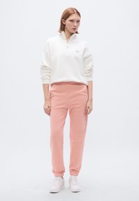 Sweatshirt branca com fecho de correr e calças de fato de treino cor de coral, com punhos e cintura elásticos. Modelo estávado de ténis brancos sobre um fundo neutro.