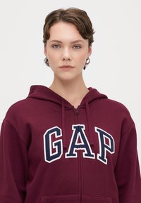 Sudadera con cremallera color burdeos hecha de algodón, que presenta un gran logotipo blanco "GAP" en el pecho y cordones en la capucha.