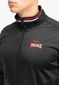 Zwarte zip-up jas met een hoge kraag, voorzien van rode en witte gestreepte accenten en een geborduurd logo op de borst. Glad textuur.