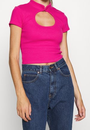 Vrouw draagt een felroze cropped top met korte mouwen en een uitsnijding aan de voorkant, gecombineerd met een hoge taille blauwe spijkerbroek, tegen een witte achtergrond.