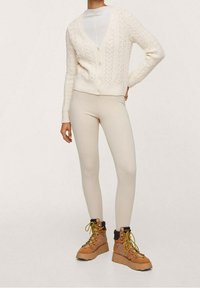 Osoba ubrana w kremowy kardigan w warkocz, biały dopasowany top, beżowe legginsy i brązowe sznurowane buty trekkingowe z grubą podeszwą, stojąca z ręką na biodrze.