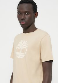 Μπεζ βαμβακερό T-shirt με μεγάλο λευκό λογότυπο δέντρου μπροστά. Κοντά μανίκια και στρογγυλή λαιμόκοψη με απαλότητα υφής.