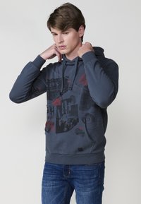 Felpa grigia scura con stampa grafica in rosso e nero, dotata di cappuccio con cordino e tasca a marsupio, abbinata a jeans blu.